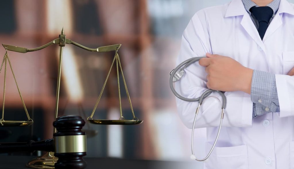 A physician holding a stethoscope stands next to judicial scales and a gavel amidst a medical malpractice lawsuit