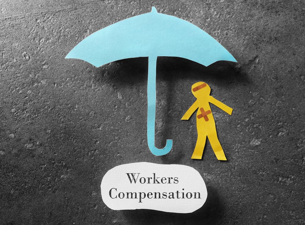 A paper art cutout of an injured stick figure under an umbrella with the words Worker's Compensation underneath